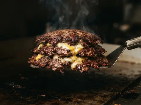 Concept imagery: three stacked smash patties with cheese oozing between each layer under a spatula